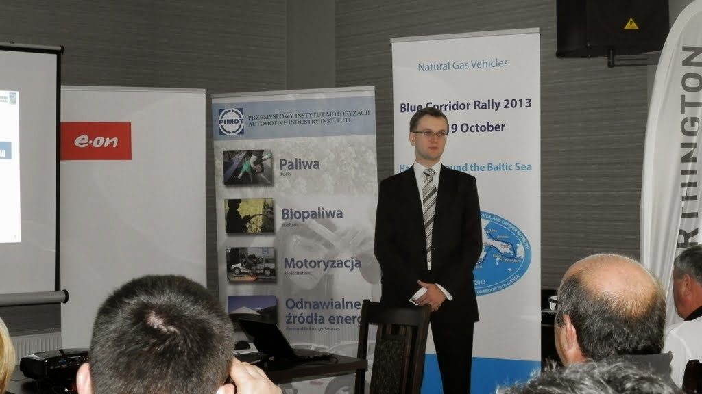 Mężczyzna w garniturze przemawia na tle rollupu Blue Corridor Rally 2013, konferencja Natural Gas Vehicles