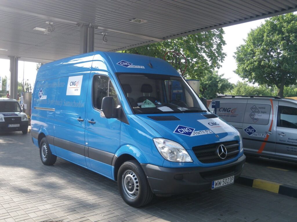 Niebieski Mercedes-Benz Sprinter z naklejkami CNG.auto.pl na stacji tankowania gazu ziemnego, w tle inny pojazd z oznaczeniem CNG