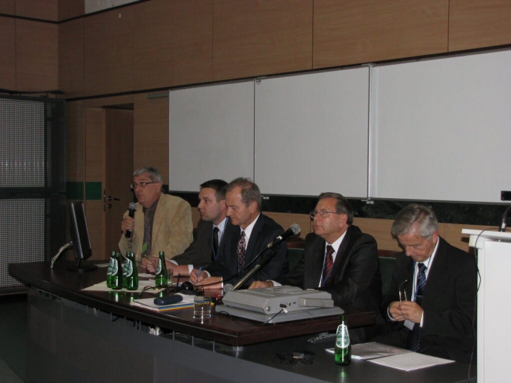 Pięciu mężczyzn w garniturach na sali konferencyjnej za stołem z butelkami wody i mikrofonami